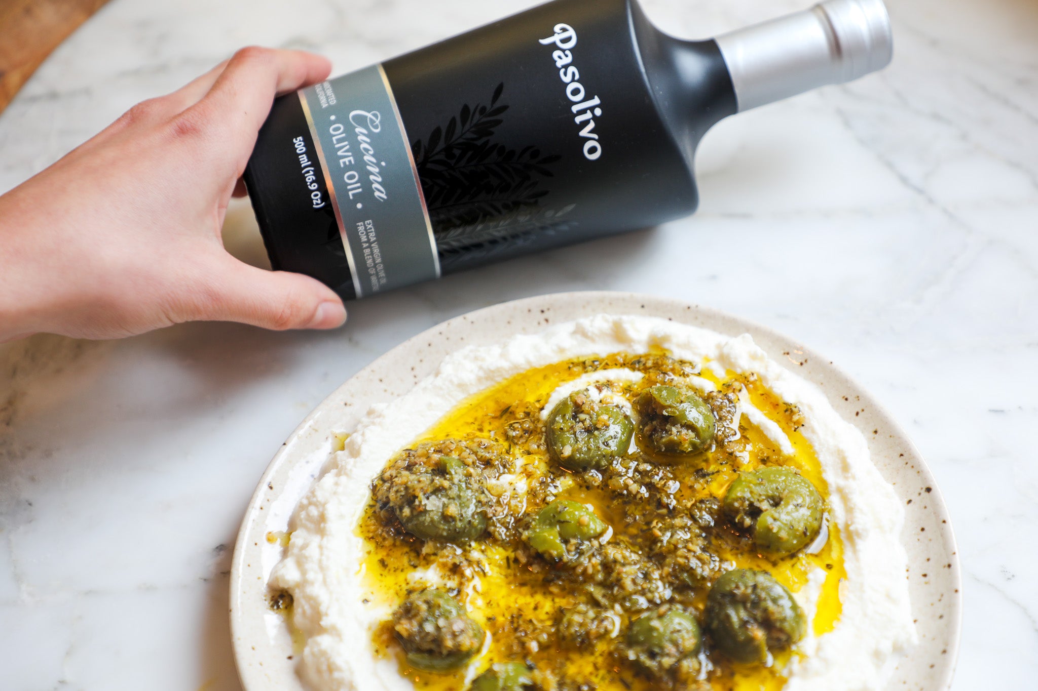 Person drizzling Pasolivo Cucina extra virgin olive oil from a bottle onto a dish with olives and cheese.
