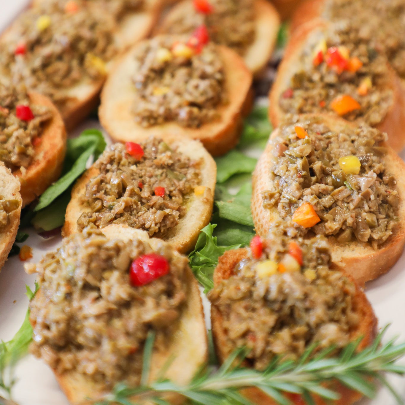 Platter of appetizers with crostini topped with green olive tapenade and garnished with herbs.