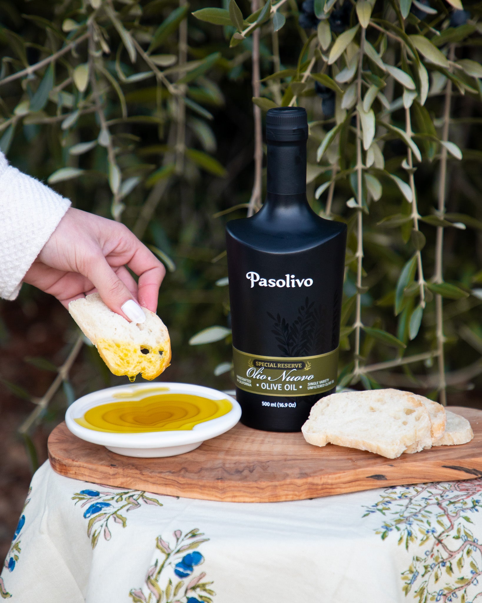Person dipping fresh bread into olive oil next to a bottle of Pasolivo Olio Nuovo.