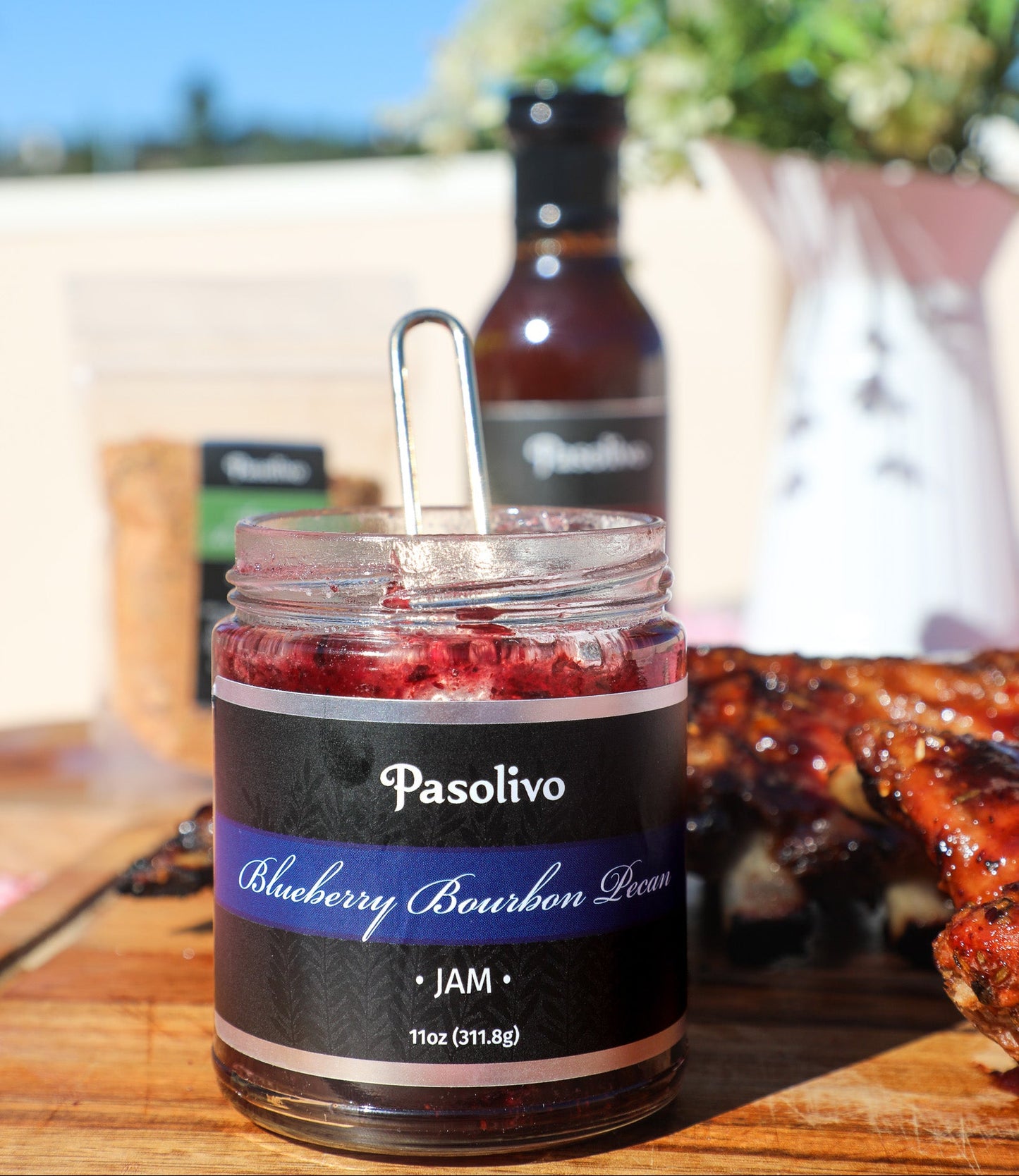 Jar of Pasolivo Blueberry Bourbon Pecan Jam with a bottle of jam in the background, on a wooden surface with a blurred outdoor setting.