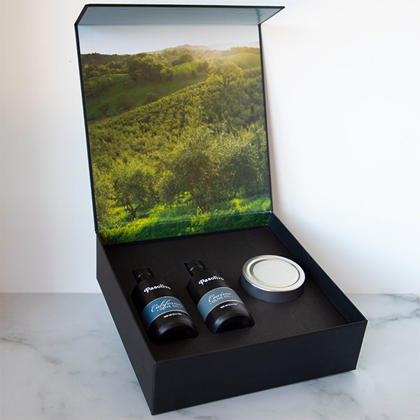 A black gift box containing two bottles of olive oil and a small jar, with a picturesque green and brown landscape image on the lid.