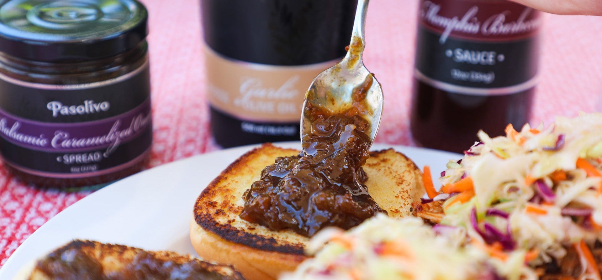 Person spooning Balsamic Caramelized Onion Spread onto BBQ Chicken Sandwiches with Sweet Habanero Coleslaw, surrounded by Garlic Olive Oil and Memphis BBQ Sauce bottles on a red cookout tablecloth.