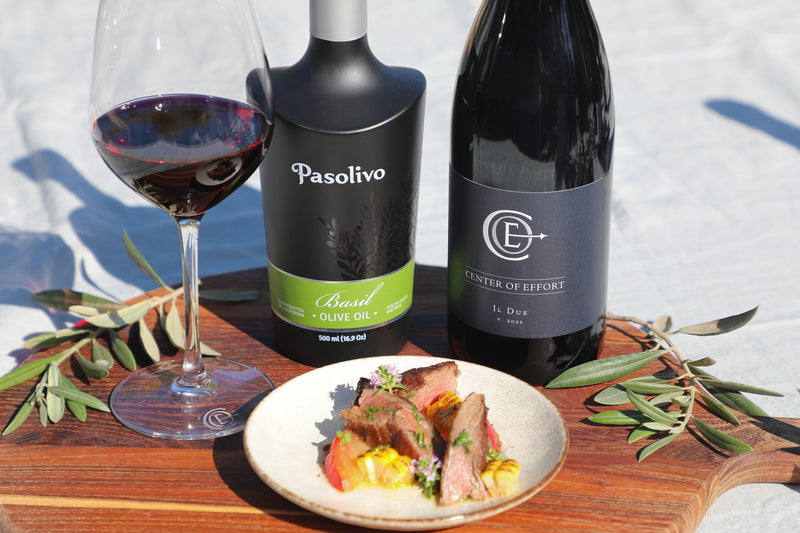 Pasolivo Basil Olive Oil, Center of Effort Il Due, and a glass of red wine arranged on a wooden board with a plated meal and olive branches, set outdoors on a white fabric surface.