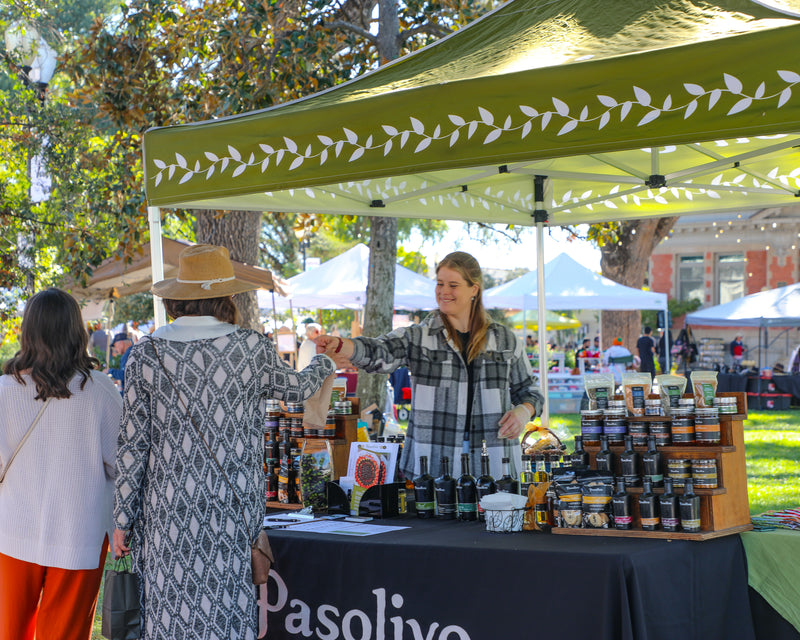 Person at a Pasolivo stand at an outdoor market with products and people around.