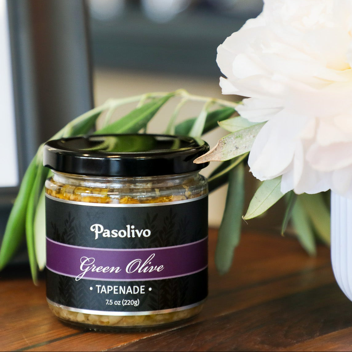 Jar of Pasolivo Green Olive Tapenade on a table with flowers in the background