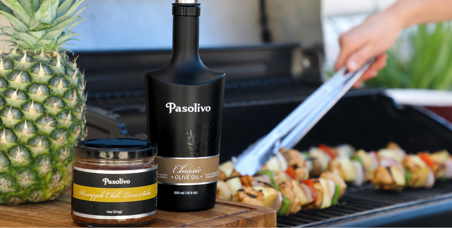 Pasolivo extra virgin olive oil bottle and jar on a cutting board with a grill and skewers in the background