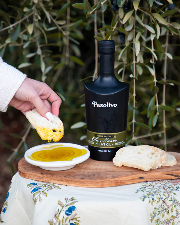 Person dipping fresh bread into olive oil next to a bottle of Pasolivo Olio Nuovo.