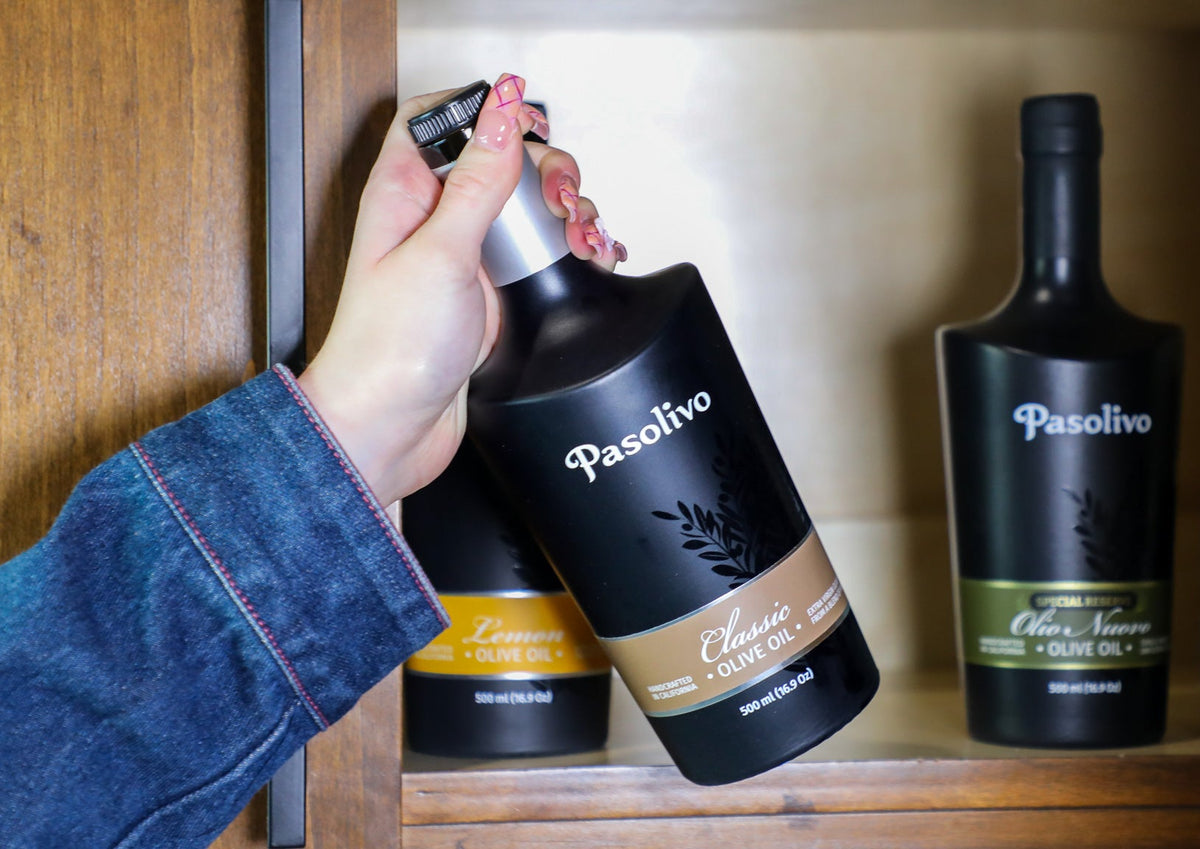 Hand holding a bottle of Pasolivo olive oil on a wooden shelf with other bottles.