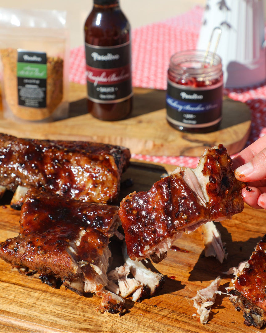 Barbecued ribs on a cutting board with sauce bottles and seasoning in the background.