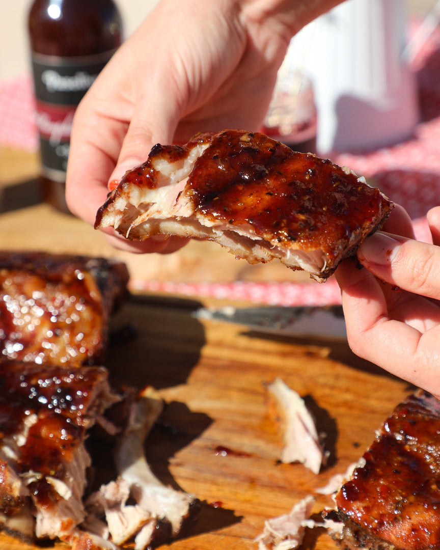 Person holding a piece of barbecue ribs on a wooden cutting board with more ribs and a bottle in the background.