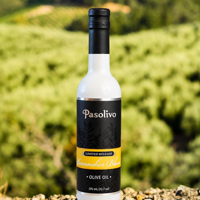 A bottle of Pasolivo Sommelier Blend extra virgin olive oil stands on a rocky surface with a blurred green olive grove background.