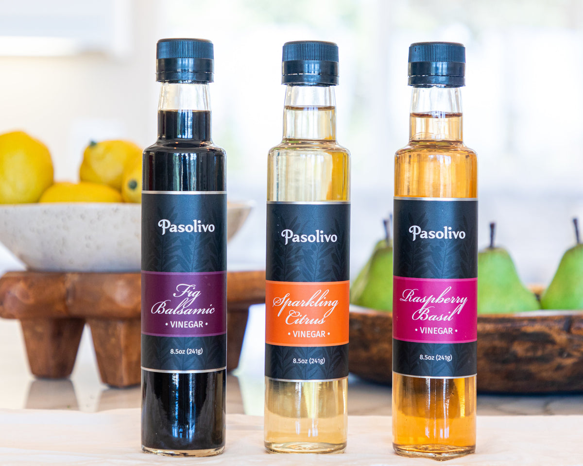 Three bottles of Pasolivo balsamic, champagne, and fruit vinegar on a kitchen counter with fruits in the background